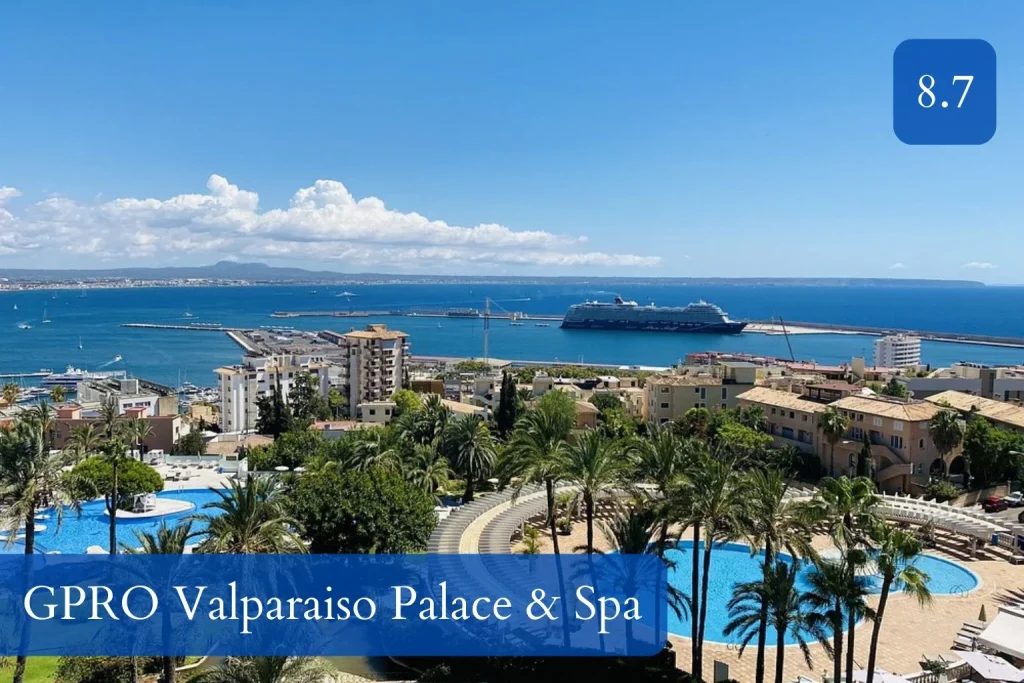 Elevated view from GPRO Valparaiso Palace & Spa featuring outdoor swimming pools, palm trees, landscaped gardens, Palma Bay, cruise ships, and the Mediterranean coastline in Bonanova, Mallorca