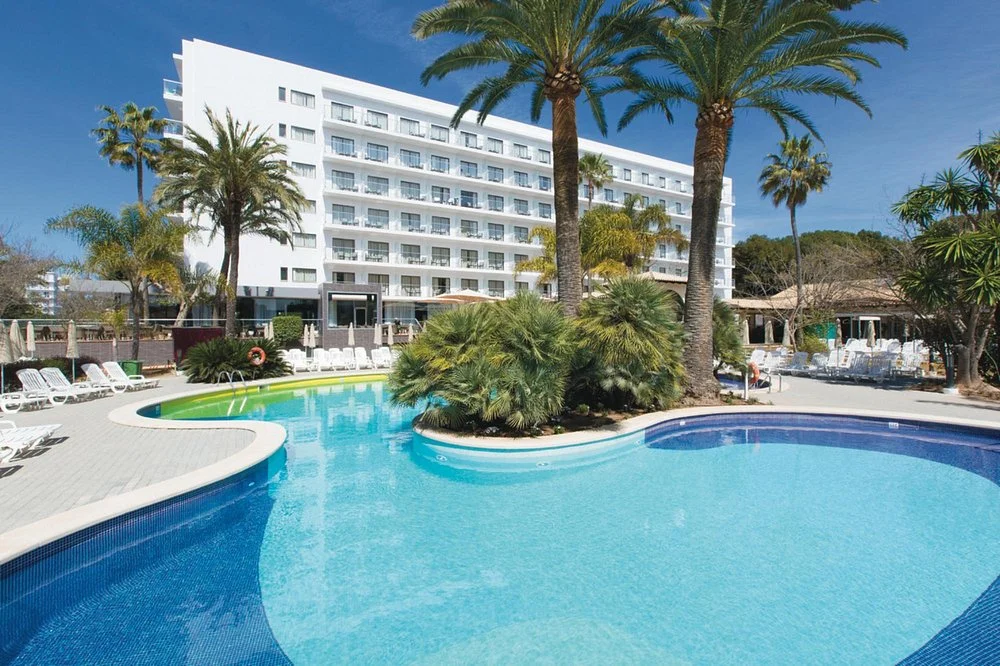 Hotel Riu Bravo in Playa de Palma with a large outdoor swimming pool, palm trees, sun loungers, and modern hotel building.