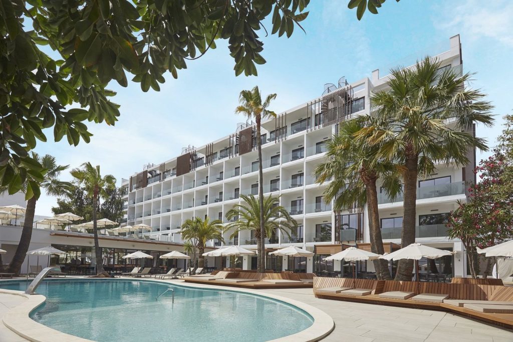 Curved outdoor pool with wooden sun loungers, white parasols, tropical landscaping, and the modern adults-only Alcudia Port Suites hotel building in Port d'Alcúdia, Mallorca