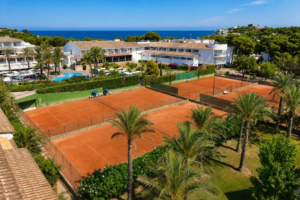 Clay tennis courts at Beach Club Font de Sa Cala on Mallorca's northeast coast with resort buildings, palm trees, and Mediterranean Sea views.