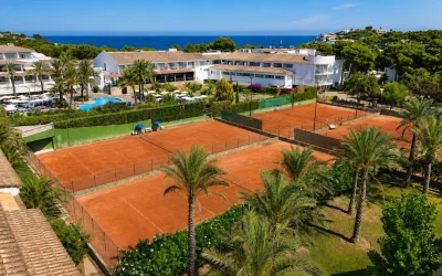 Clay tennis courts at Beach Club Font de Sa Cala on Mallorca's northeast coast with resort buildings, palm trees, and Mediterranean Sea views.