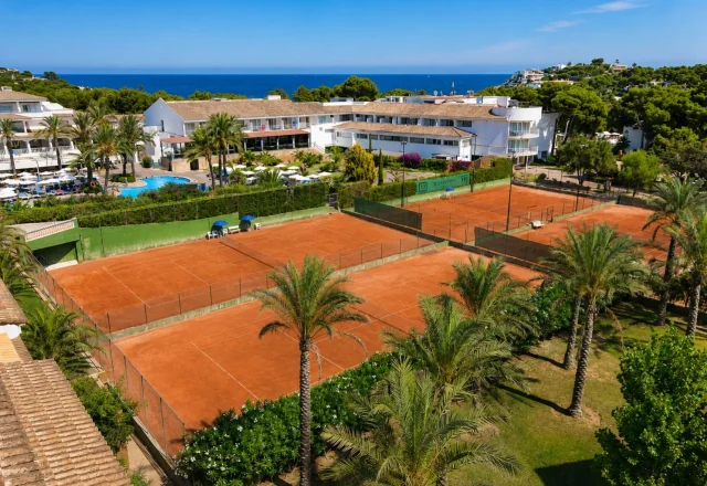 Clay tennis courts at Beach Club Font de Sa Cala on Mallorca's northeast coast with resort buildings, palm trees, and Mediterranean Sea views.