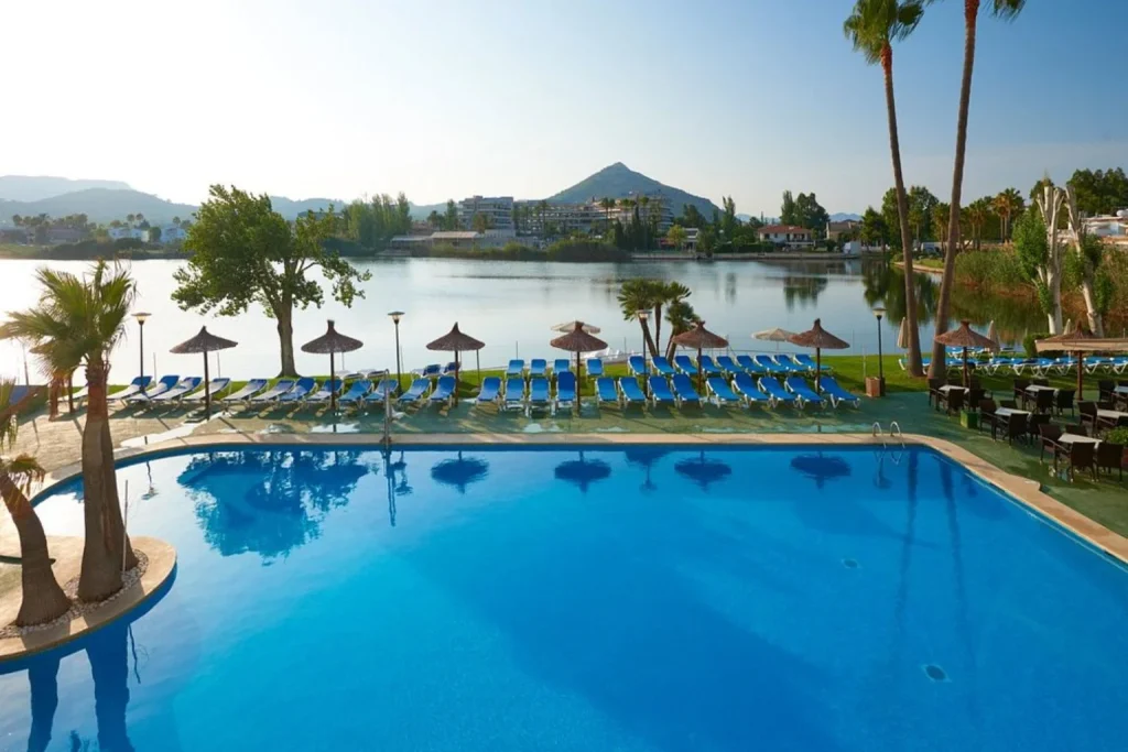 Outdoor swimming pool with spacious sun terrace, loungers, parasols, lakeside setting, and mountain views at Grupotel Amapola near Playa de Muro, Mallorca