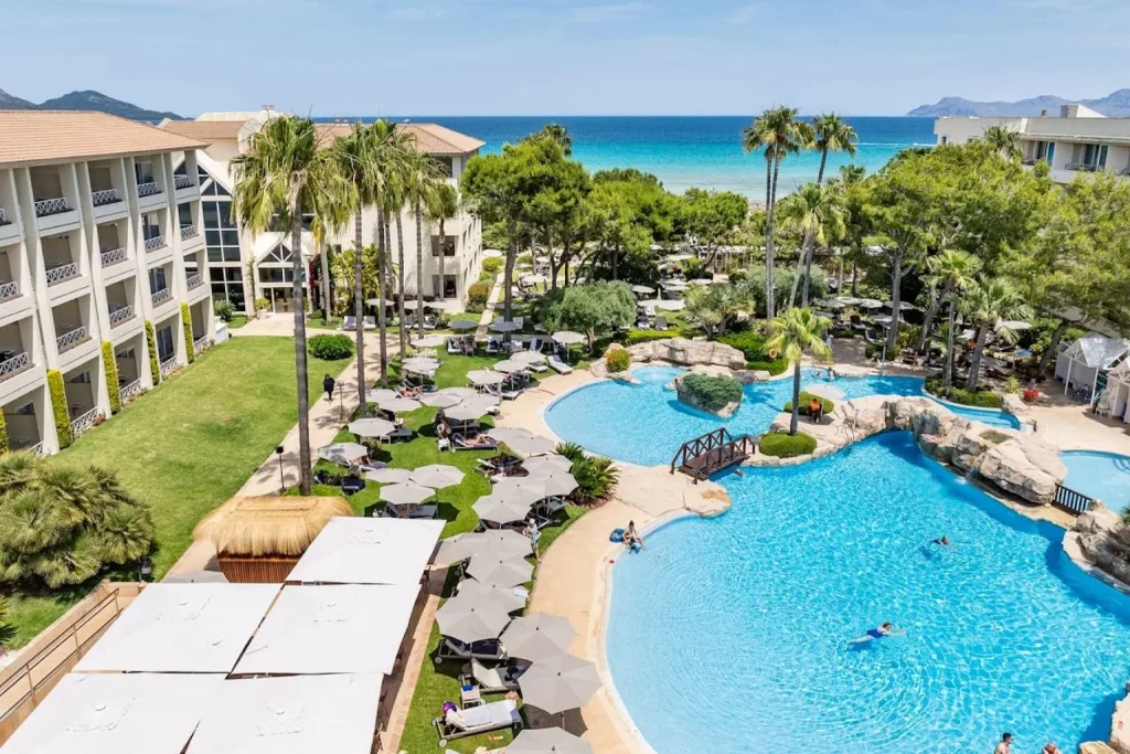 Lagoon-style outdoor pools with rock features, a small wooden bridge, sun loungers, landscaped gardens, and Mediterranean Sea views at Grupotel Parc Natural & Spa in Playa de Muro, Mallorca