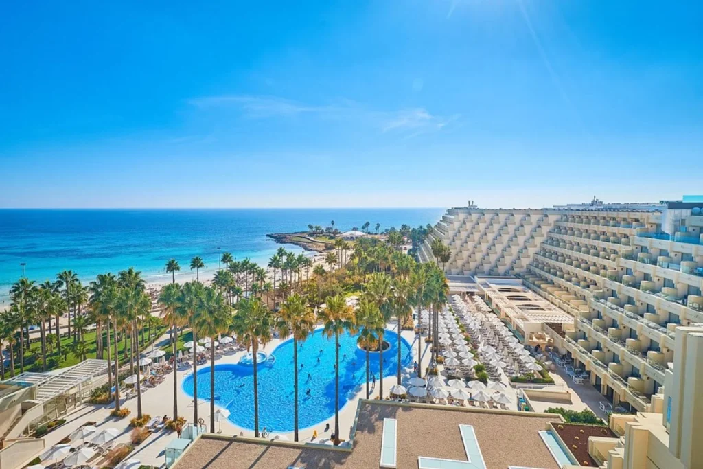 Lagoon-style outdoor swimming pool with island whirlpool, sun loungers, beachfront setting, Mediterranean Sea views, and modern resort buildings at Hipotels Mediterráneo Hotel in Sa Coma, Mallorca