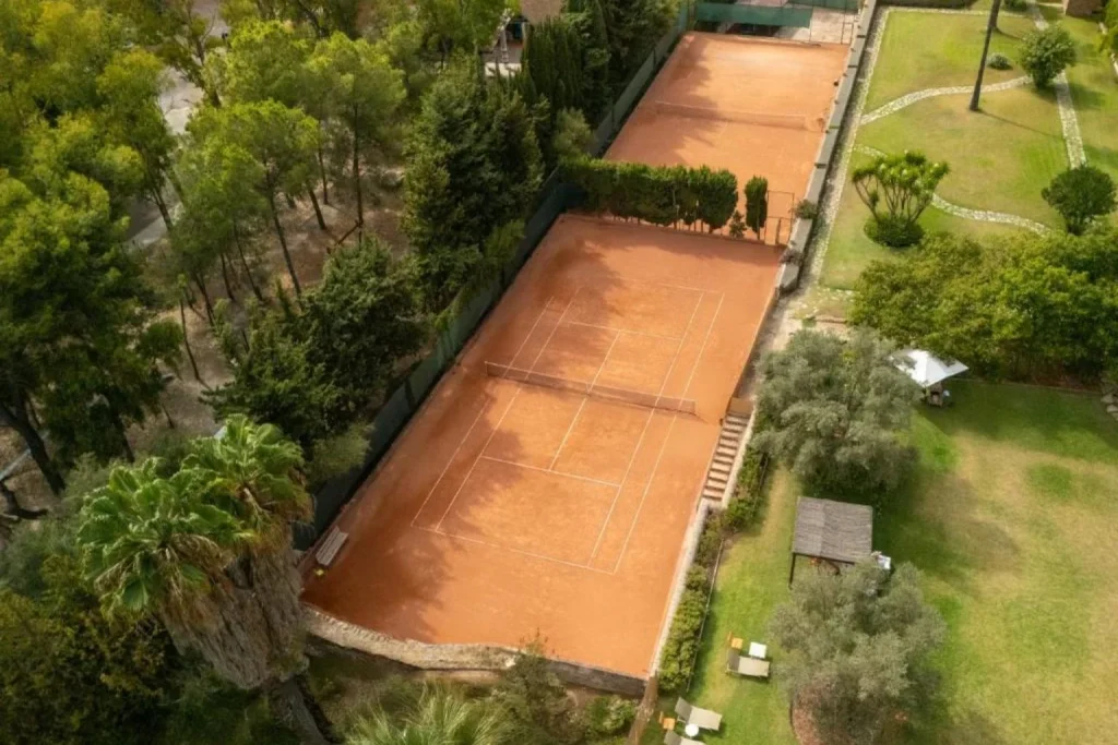 Aerial view of clay tennis courts surrounded by gardens and trees at Hotel Es Port in Sóller.