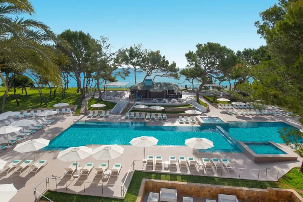 Large outdoor swimming pool with sun loungers, pine trees, and direct beachfront setting at Iberostar Selection Playa de Muro Village in Playa de Muro, Mallorca.