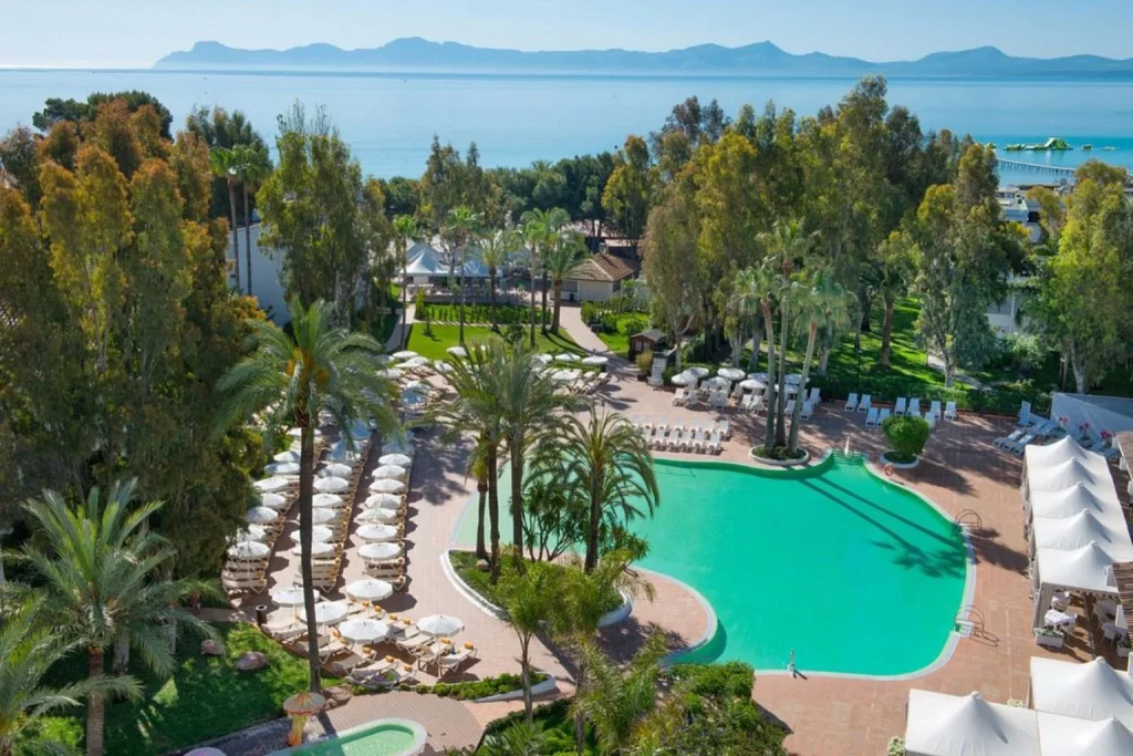 Lagoon-style outdoor swimming pool with island planters, sun loungers, parasols, landscaped gardens, and Mediterranean Sea views at Iberostar Waves Ciudad Blanca in Alcudia, Mallorca