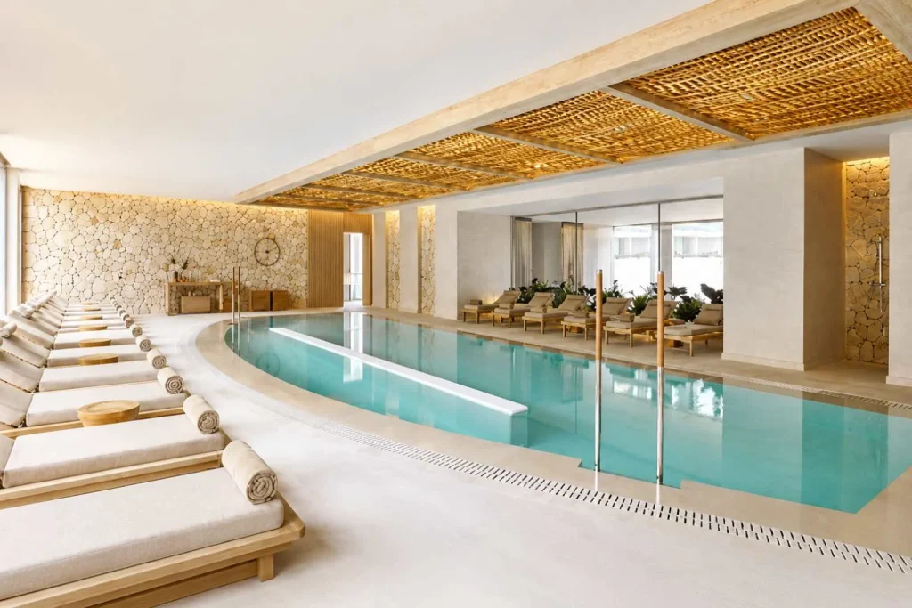 Indoor spa pool with relaxation loungers, natural stone walls, and wooden ceiling at Ikos Porto Petro in Porto Petro, Mallorca.