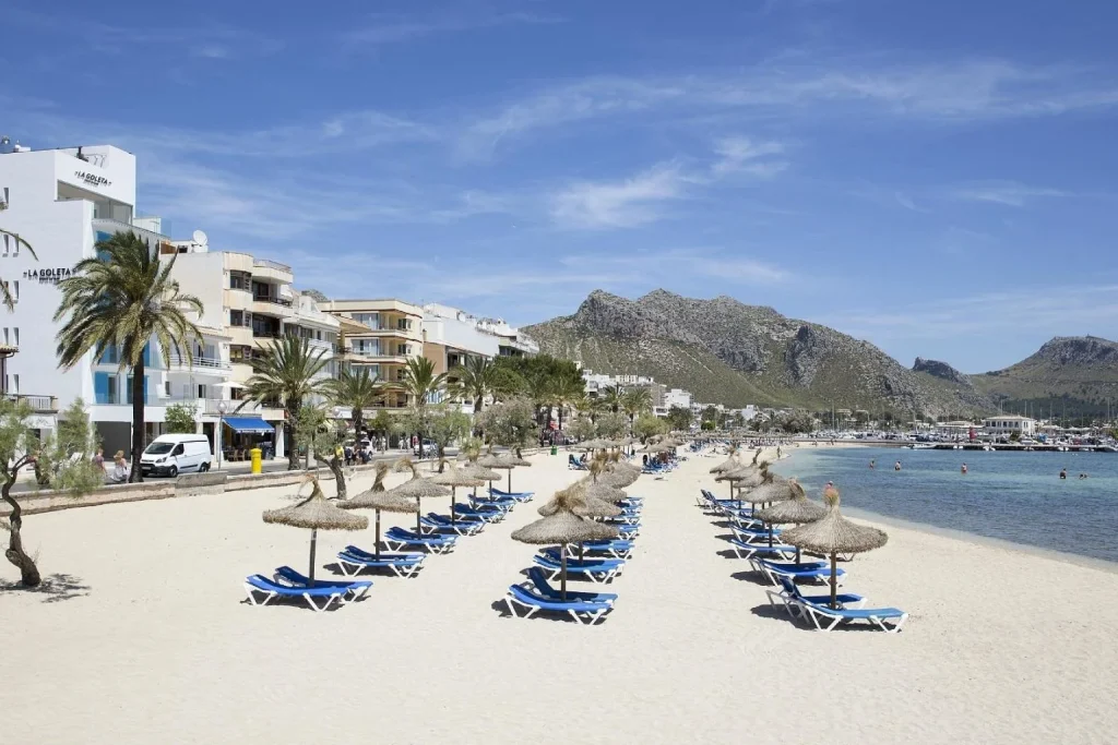 Beachfront view with a sandy beach, marina, and mountain backdrop at La Goleta Hotel de Mar in Port de Pollença, Mallorca.