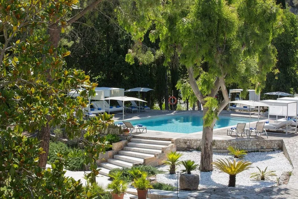 Outdoor swimming pool surrounded by Mediterranean gardens, cabanas, and mature trees at L’Hermitage Hotel & Spa in Orient, Mallorca.