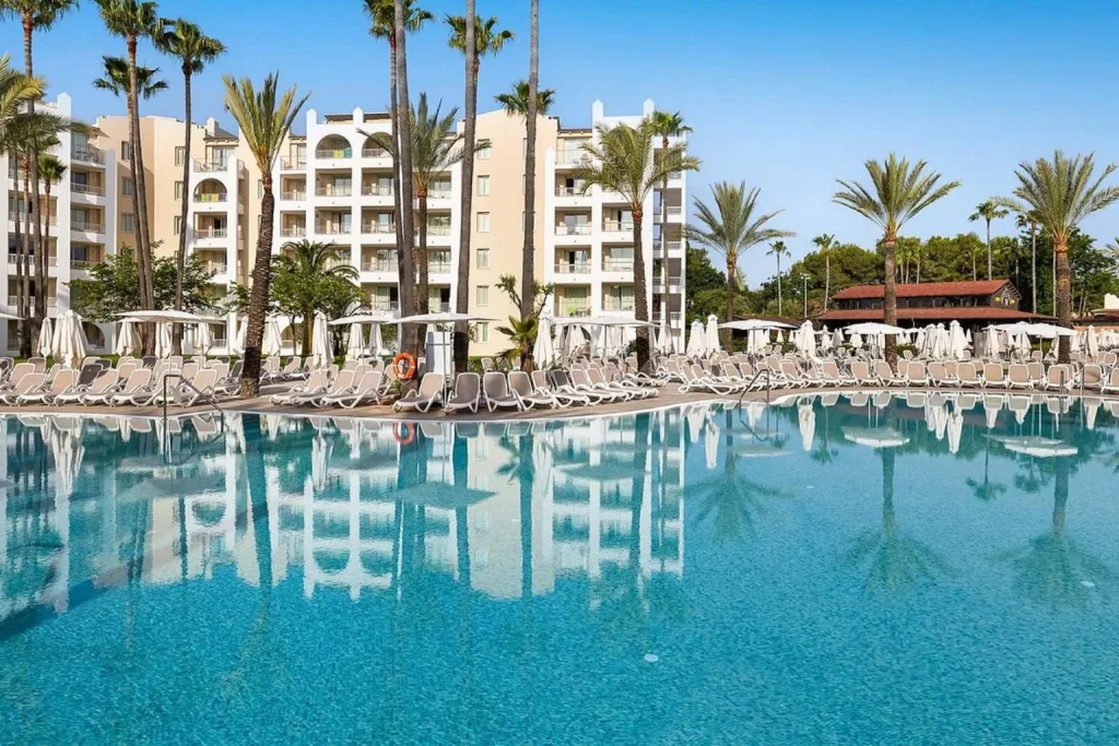 Large outdoor swimming pool surrounded by palm trees at Protur Safari Park Aparthotel in Sa Coma, Majorca.